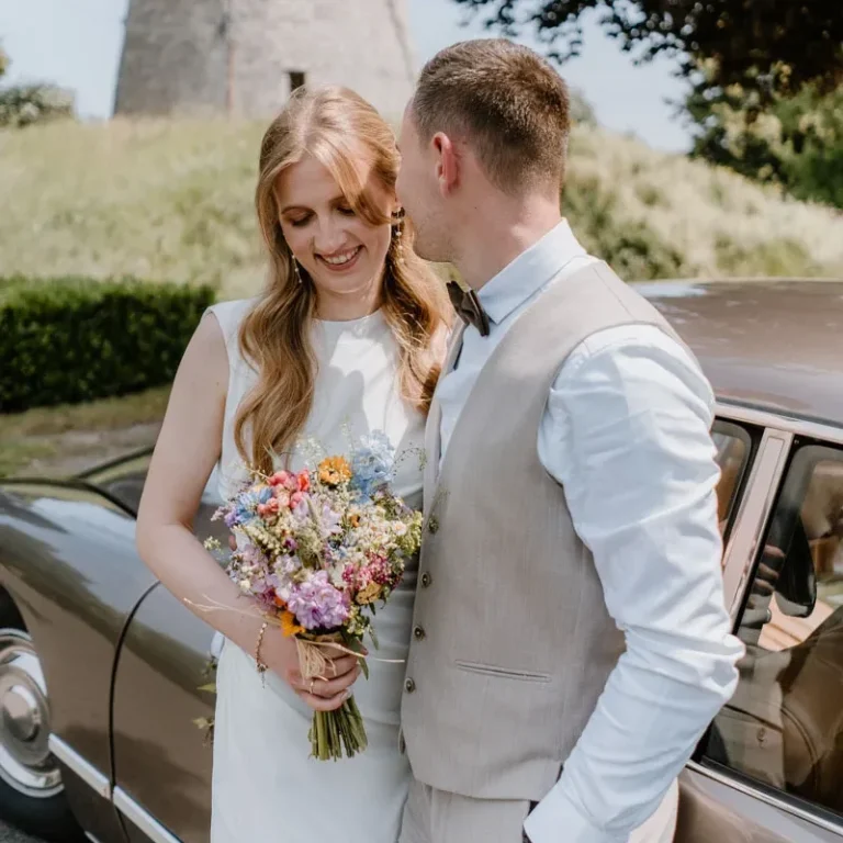 Brautpaarshooting mit Oldtimer bei Mühle Ennigerloh bei Münster Ein Brautpaar steht lächelnd neben einem Oldtimer und hält einen bunten Blumenstrauß.