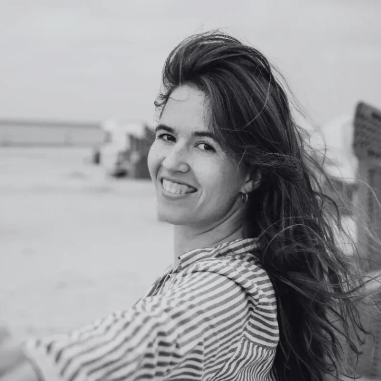 Hochzeitsfotografin Fabi aus Münster Lächelnde Frau mit langen, windbewegten Haaren vor einem Strandhintergrund.